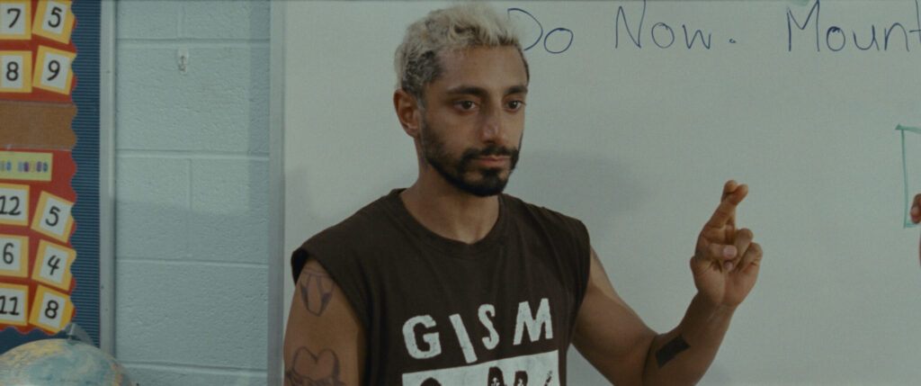 Image description: Main character Ruben, played by Riz Ahmed, stands in front of a white board in a classroom and holds up his left hand to sign the letter "r" in American Sign Language. He has a beard and short bleached hair. He is wearing a black t-shirt with the band name GISM and has numerous tattoos visible on his arms.